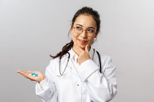 Healthcare, medicine and pharmacy concept. Portrait of thoughtful, pleased female asian doctor, smirk and look up, thinking satisfied, holding pills, invent new drugs to cure sick people