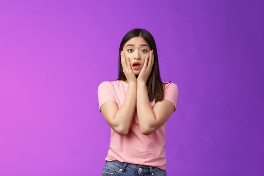 Concerned shocked timid insecure asian woman, feel pitty, shame hearing shocking news, grab face sorry for friend, gasping, opened mouth upset, sighing distressed, stand purple background