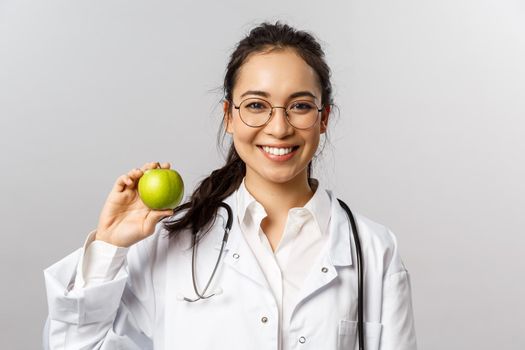 An apple a day keeps doctors away. Portrait of optimistic cute asian female doc in white coat, advice eating healthy food, more vitamins, holding fruit and smiling camera, grey background