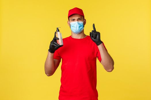 Covid-19, delivery orders, shopping, contactless handling and social distancing concept. Serious-looking courier in medical mask and gloves, red uniform, tell to use hand sanitizer, rule number one