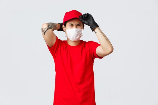 Reluctant asian delivery guy put on red cap, frowning and grimacing displeased, wear protective gloves and medical mask, unwilling carry heavy box. Courier complaining hard work