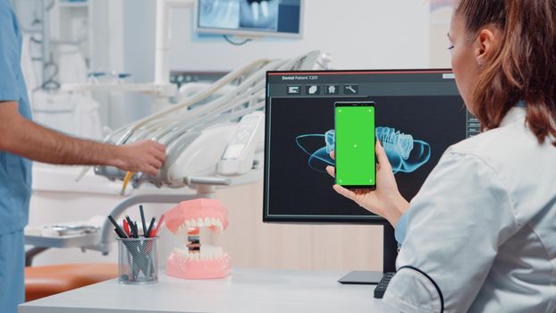 Dentist vertically holding smartphone with green screen while looking at teeth scan on computer in office. Teethcare specialist using mobile phone with chroma key for isolated background