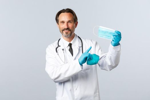 Covid-19, preventing virus, healthcare workers and vaccination concept. Smiling male doctor in white coat and gloves, preventive measures social distancing, hold respirator and point medical mask