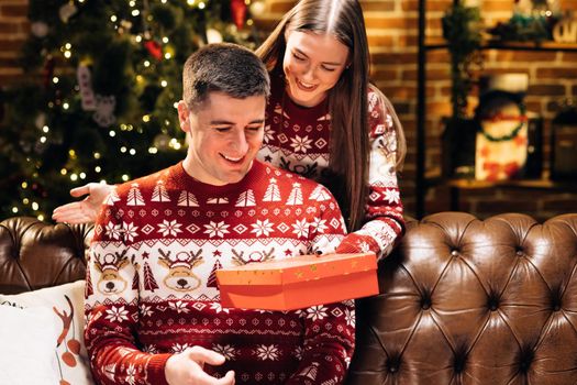 Young 30s woman covering eyes of smiling curious husband, giving wrapped box with Christmas gift near decorated festive tree, New Year winter holidays family celebration. Holiday miracle