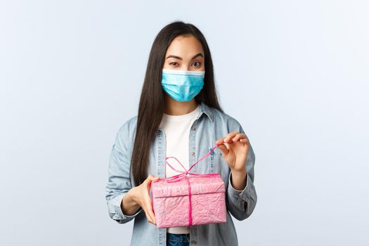 Social distancing lifestyle, covid-19 pandemic, celebrating holidays during coronavirus concept. Intrigued happy asian woman in medical mask unwrap gift box wonder what inside