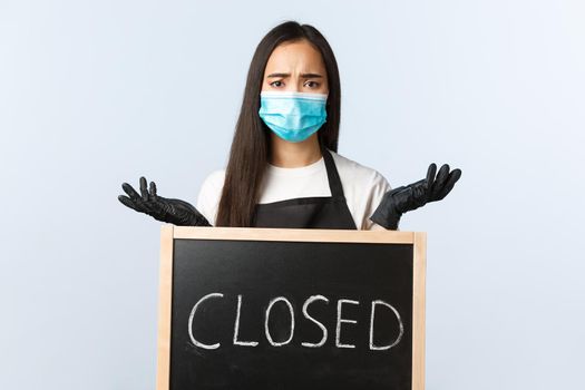 Small business, covid-19 pandemic, preventing virus and employees concept. Complicated and confused asian employee, barista in medical mask shrugging over closed sign
