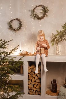 A girl is playing on Christmas Eve in a beautiful house decorated for the New Year holidays. Scandinavian-style interior with live fir trees and a wooden staircase.