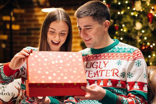 Portrait of cheerful young woman opening xmas gift box with excited surprised face feeling happy on New Year's Eve sitting at home near glowing christmas tree. Xmas concept. Holiday miracle