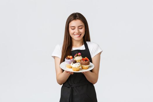 Grocery store employees, small business and coffee shops concept. Cheerful silly waitress want have bite of delicious cupcakes. Barista biting lip tempting to taste new desserts, white background