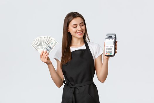 Grocery store employees, small business and coffee shops concept. Smiling pleased barista in black apron looking at payment terminal satisfied as showing cash, prefer POS paying
