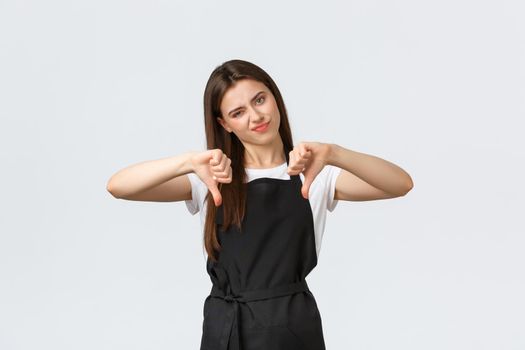 Grocery store employees, small business and coffee shops concept. Skeptical and disappointed waitress in black apron smirk displeased and showing thumbs-down, white background