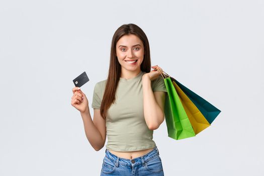 Shopping mall, lifestyle and fashion concept. Excited smiling young woman biting lip, tempt to waste all money on credit card, holding bags with clothes and looking pleased, white background