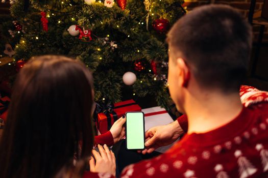 Couple using smartphone for christmas video call with green mock up screen. Christmas, festivity and communication technology. Hands holding a smart phone with green mock up screen