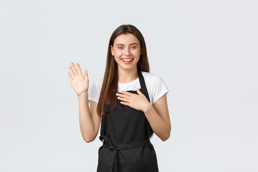 Grocery store employees, small business and coffee shops concept. Sincere smiling cute female barista swear with one raised hand and another on heart. Pretty saleswoman tell truth, make promise