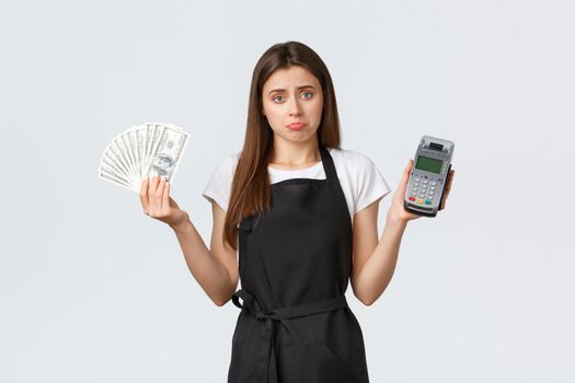 Grocery store employees, small business and coffee shops concept. Indecisive cute shrugging waitress pouting unsure and showing cash with POS terminal, standing white background