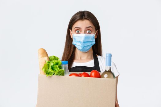 Covid-19 social distancing, delivery and grocery shopping during coronavirus concept. Young pleasant saleswoman, cashier in medical mask handling over groceries to customer