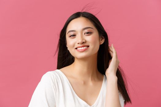 Beauty products advertisement, haircare and women fashion concept. Close-up of sensual beautiful korean woman smiling broadly with white teeth, touching haircut gently, stand pink background
