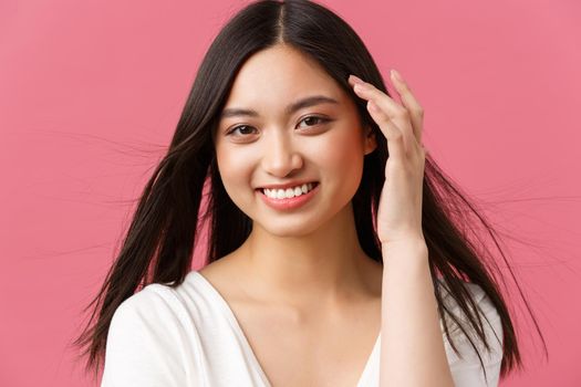 Beauty salon, haircare and skincare products advertisement concept. Gorgeous asian woman 20s, gently touching hair and smiling with shy romantic expression, standing pink background