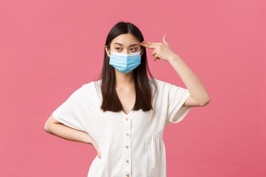 Covid-19, social distancing, virus and lifestyle concept. Annoyed and distressed asian female student in medical mask, showing fake gun over temple as killing herself from boredom or annoyance
