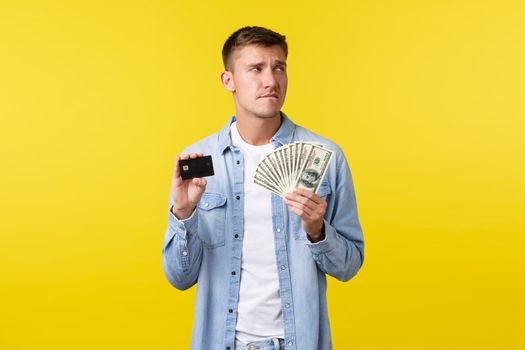 Thoughtful and indecisive, worried handsome man in casual clothes, looking perplexed away and biting lip, tempting to buy something expensive, showing credit card with money