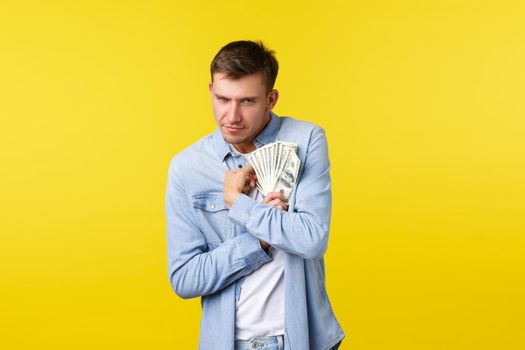 Investment, shopping and finance concept. Greedy funny blond guy hugging money and looking at camera, showing intense desire to keep cash, unwilling share, standing yellow background