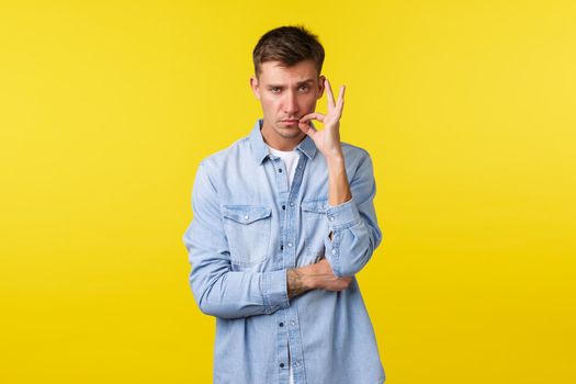 Portrait of serious-looking blond caucasian man zipping his mouth on lock, swear keep secret and stay silent, seal lips with promise, standing determined over yellow background