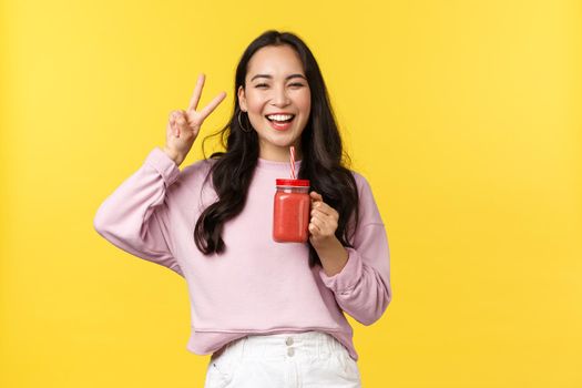 People emotions, lifestyle leisure and beauty concept. Happy upbeat asian 20s girl showing peace sign and smiling delighted as enjoying summer drink, holding smoothie, yellow background