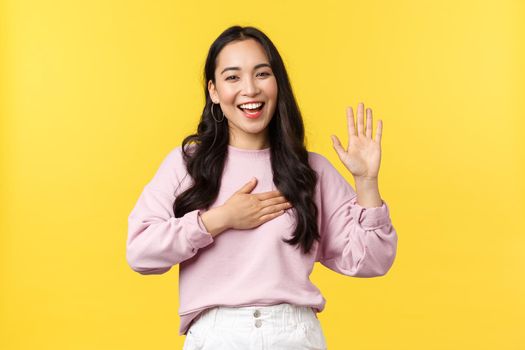 Lifestyle, emotions and advertisement concept. Honest and sincere happy smiling asian woman promise tell truth, swear on her heart and raising one hand, standing yellow background