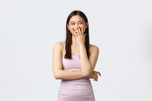 Luxury women, party and holidays concept. Coquettish and carefree beautiful asian girl in evening prom dress, laughing, cover mouth shy and smiling, looking right, white background