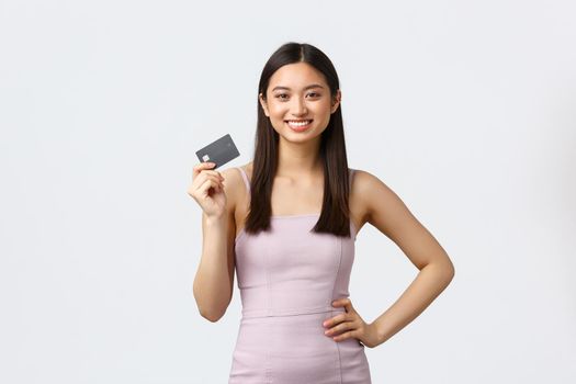 Luxury women, party and holidays concept. Gorgeous young asian female in evening dress, showing credit card, ready go shopping to mall, invest money, smiling satisfied, white background