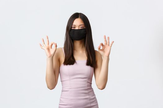 Covid-19, social distancing quarantine and leisure concept. Elegant pretty asian woman in evening dress, prom celebration, show okay signs and wear face mask to protect from coronavirus at party