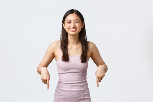 Luxury women, party and holidays concept. Excited happy asian girl attend prom in evening dress, smiling broadly with rejoice, pointing fingers down, demonstrate banner, white background