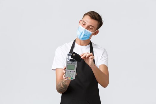 Coronavirus, social distancing in cafes and restaurants, business during pandemic concept. Handsome man in medical mask showing credit card and POS terminal, using contactless way of payment