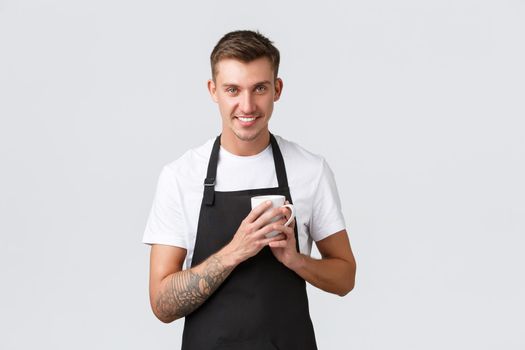 Small business, coffee shop, cafe and restaurants concept. Handsome barista enjoying making coffee, holding mug near heart and smiling cheeky camera, white background