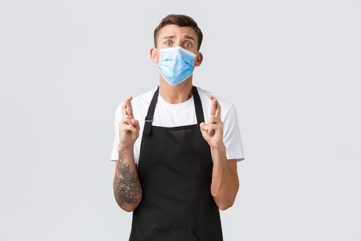 Coronavirus, social distancing in cafes and restaurants, business during pandemic concept. Desperate and worried salesman, owner of store cross fingers good luck, wearing medical mask and praying