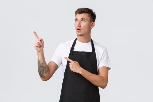 Small business owners, coffee shop and staff concept. Worried and upset gloomy cafe employee, restaurant waiter or barista in apron, pointing looking upper left corner with regret and disappointment