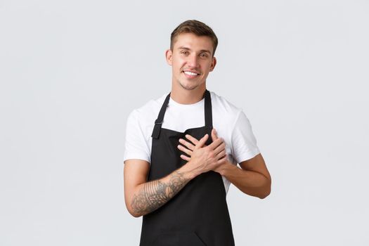 Small business owners, coffee shop and staff concept. Happy handsome blond guy in black apron, barista or waiter holding hand on heart and smiling, cherish cafe guests, love every customer