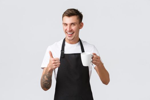 Small business, coffee shop, cafe and restaurants concept. Friendly handsome waiter, barista selling drink, handing cappuccino in mug guest, smiling happy, white background, recommend beverage