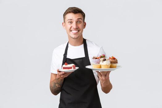 Small retail business, coffee shops and bakery concept. Friendly handsome barista, waiter in black apron serving tables, holding delicious desserts cakes and cupcakes, white background
