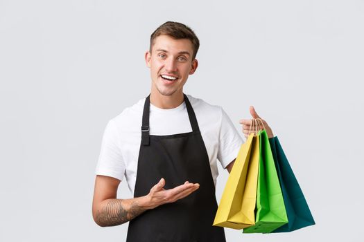Retail store, shopping and employees concept. Friendly and polite handsome salesman, shop assistant handing over eco-bags with client purchased items, white background