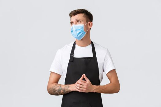 Coronavirus, social distancing in cafes and restaurants, business during pandemic concept. Disappointed and sad barista, salesman in apron and medical mask looking left displeased and upset