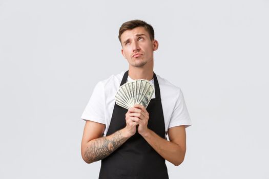 Small retail business, payments and employees concept. Thoughtful handsome barista, salesman in black apron, looking upper left corner and holding money, thinking what buy on paycheck