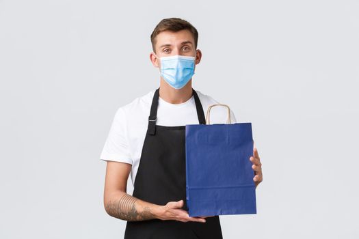 Retail store, shopping during covid-19 and social distancing concept. Friendly polite salesman, barista in medical mask and black apron, put purchased item in eco-bag, standing white background