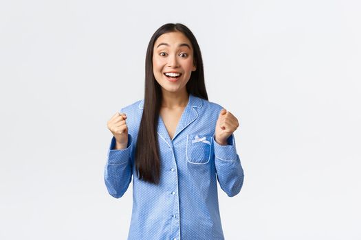 Hopeful excited asian girl in blue pajamas clench fists and looking rejoice camera, smiling as awaiting great news, feeling enthusiastic and upbeat as standing over white background