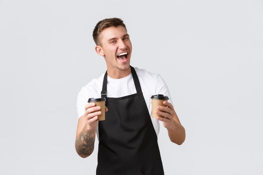 Small business, coffee shop, cafe and restaurants concept. Enthusiastic happy barista in black apron holding two paper cups with coffee, selling and giving order to clients, laughing joyfully