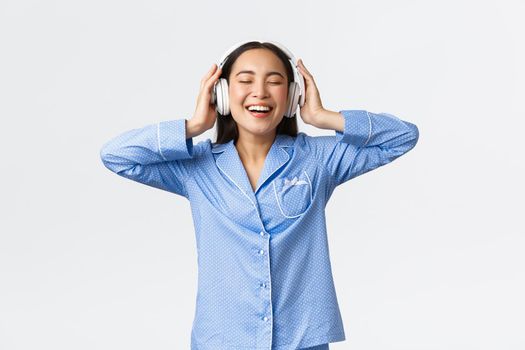 Home leisure, weekends and lifestyle concept. Happy pleased asian woman in pajamas enjoying awesome sound quality in new earphones, dancing in pajamas and listening music