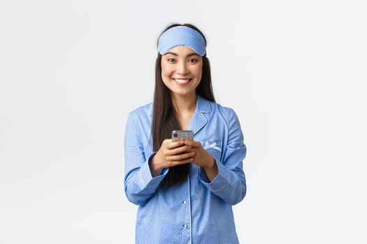 Technology, people and home leisure concept. Smiling cute asian female blogger in pajamas and sleeping mask, writing post in social media before sleep, messaging with mobile phone.
