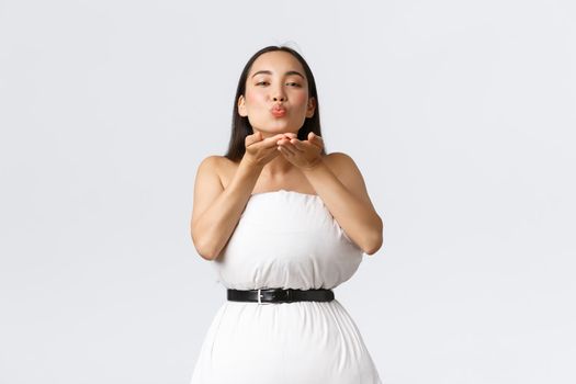Beauty, fashion and social media concept. Gorgeous asian woman making dress from pillow secured with belt around waste, blowing air kiss at camera coquettish over white background