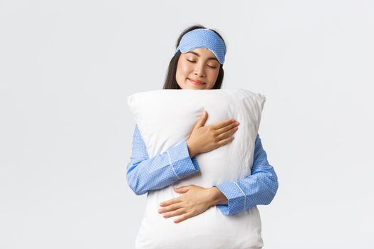 Beautiful smiling asian girl in sleeping mask and pajama, having sweet dream, hugging pillow with silly grin and closed eyes, standing over white background, unwilling wake-up