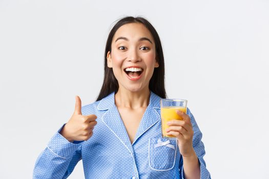 Healthy lifestyle, morning routine and people concept. Close-up of excited beautiful asian girl in pajama having habit of drinking fresh orange juice full of vitamins, showing thumbs-up